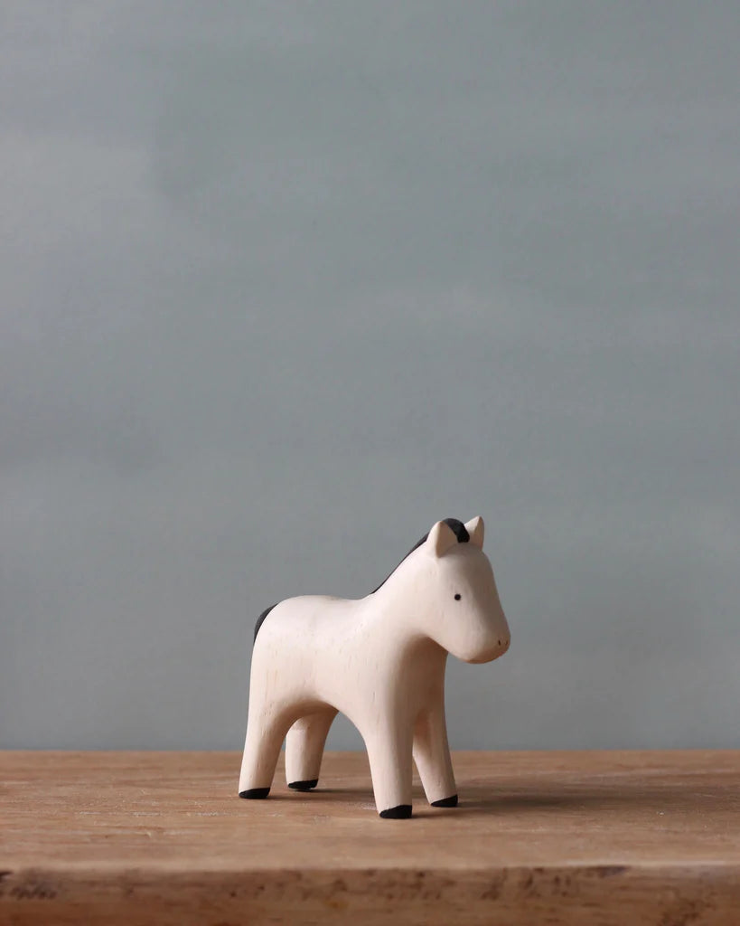 A Handmade Tiny Wooden Farm Animals - Horse figurine on a wooden surface against a soft gray background. The horse is painted white with black features.