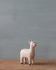 A small, hand-painted alpaca figurine from the "Handmade Tiny Wooden Farm Animals - Alpaca" collection stands on a wooden surface against a plain, light gray background. The handcrafted wooden animal features a minimalist design in light beige with black tips on its ears and feet.