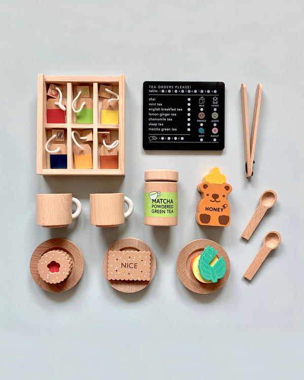 Experience the charm of the Wooden Tea Set with organic Matcha Tea, colorful teabags, two cups, a matcha canister, honey jar, tongs, dessert pieces on plates, spoons, and a tea order card—all set against a light gray background for a delightful play space.