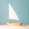 The Bumbu Handmade Wooden Sailing Boat - White, featuring a single white sail, is displayed on a light wooden surface with a plain light blue background.