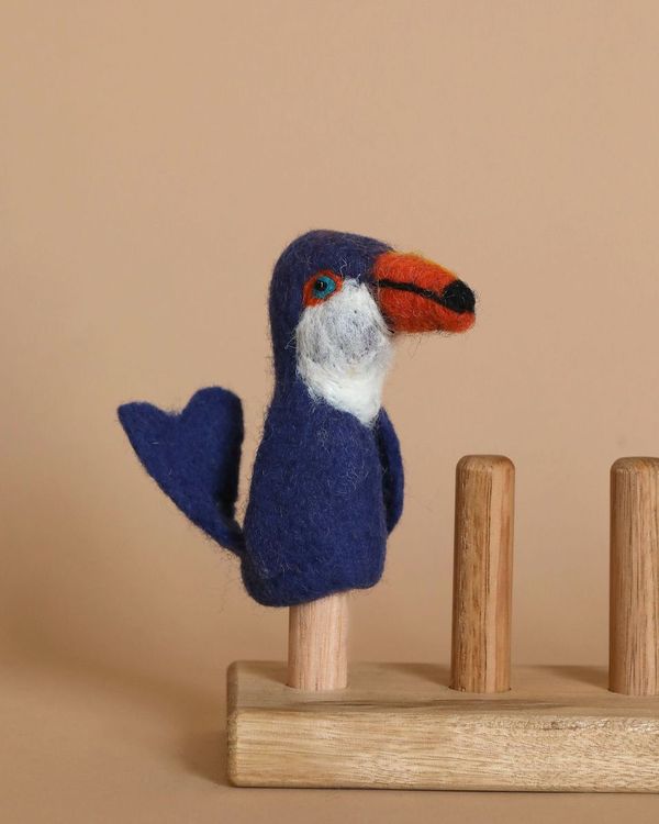 The Toucan Finger Puppet, a small, handcrafted felt bird with a blue body, white chest, and orange beak, is perched on a wooden stand against a beige background. Made from 100% wool using the needle felted technique, this charming piece boasts intricate detail and charm.