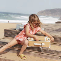 A young girl with blond hair, wearing a peach dress and yellow sandals, plays with a beige Olli Ella See-Ya Suitcase - Butterscotch on a wooden boardwalk by the beach. Waves crash in the background under a