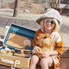 A young child in sunglasses and a wide-brimmed hat sits on a wooden boardwalk, holding a stuffed lion toy, with an open Olli Ella See-Ya Suitcase - Butterscotch beside them.