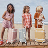 Three young children stand on a wooden boardwalk at the beach, each holding a Olli Ella See-Ya Suitcase - Butterscotch. The children appear to be ready for a journey, showcasing playful yet thoughtful expressions.
