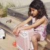A young girl sits on a wooden bench outdoors, resting her arms on a pink, polka-dotted Olli Ella See-Ya Suitcase - Pink Daisies, with an open book and a toy car nearby, appearing thoughtful.