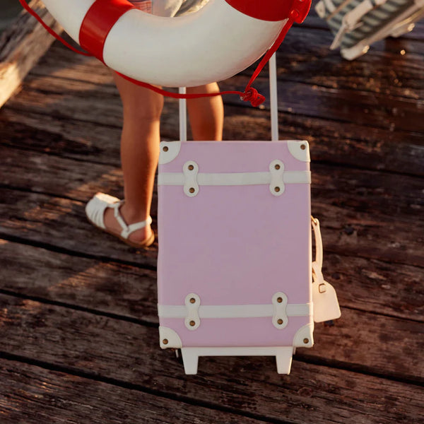A person stands on a wooden dock with the Olli Ella See-Ya Suitcase - Pink, featuring a retractable handle and wearing white sandals, while a lifebuoy with red accents is visible in the background.