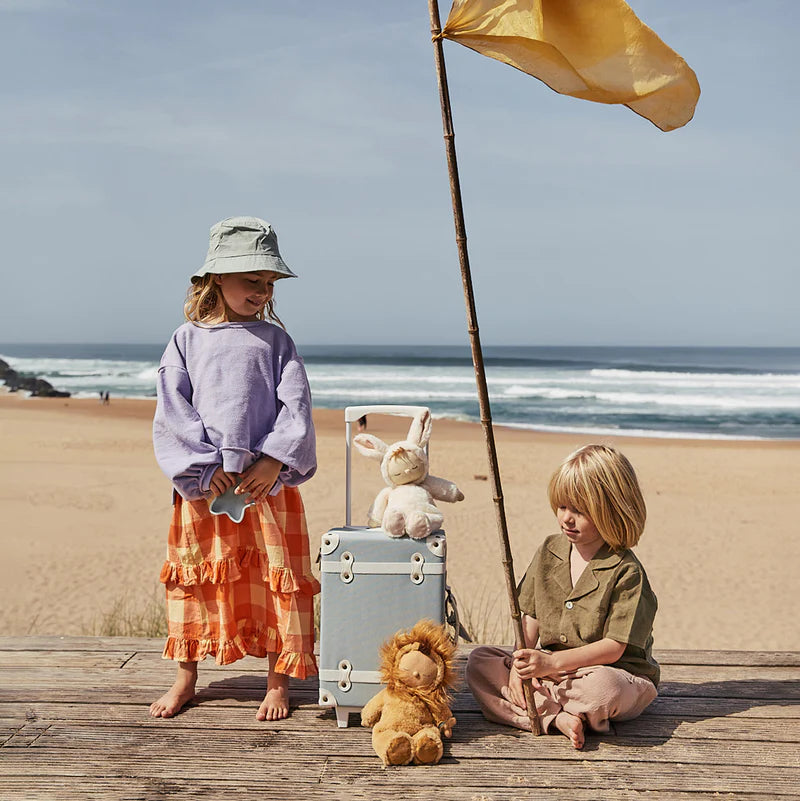 Two children dressed in casual outfits are at a beach with an Olli Ella See-Ya Suitcase - Steel Blue and toys, a yellow flag waving on a pole next to them, with the sea in the background.