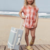 A young child in a peach plaid shirt and yellow flip-flops stands on a wooden boardwalk holding an Olli Ella See-Ya Suitcase - Steel Blue, with the sandy beach and ocean in the background.