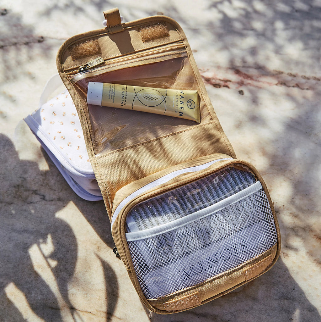 An open Olli Ella See Ya Wash Bag - Butterscotch on a sunny surface, containing sunscreen, a notepad, and other small items, displaying organized compartments.