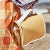 A person carrying a stylish Butterscotch Olli Ella See Ya Wash Bag by the seaside. The bag is matched with a colorful plaid shirt, and there is a sandy beach in the background.