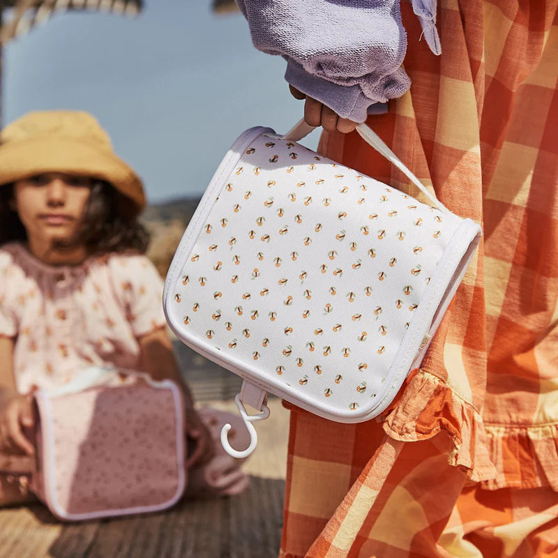 A person holding an Olli Ella See Ya Wash Bag - Leafed Mushroom, made from recycled PET, stands near a child in a floppy hat. The setting suggests a sunny outdoor scene.