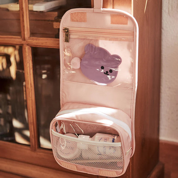 A pink Olli Ella See-Ya Wash Bag hanging organizer with multiple compartments, including a transparent section with a purple pouch and a lower mesh pocket containing various electronic devices, mounted on a wooden cabinet.