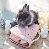 A small gray rabbit sitting on a pink, star-patterned Olli Ella See Ya Wash Bag - Pink Daisies, surrounded by beauty products and a light blue case, on a bright, sunny day.