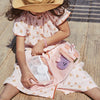 A young girl in a peach dress and sunhat sits on a wooden deck, looking into an open Olli Ella See Ya Wash Bag - Pink Daisies revealing a sandwich and a recycled plastic container.