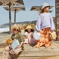 Three children in casual summer clothes and hats stand on a wooden walkway by the beach, holding colorful buckets. The background shows a sandy beach and clear skies, perfect for a day out with their Olli Ella See Ya Wash Bag - Steel Blue.