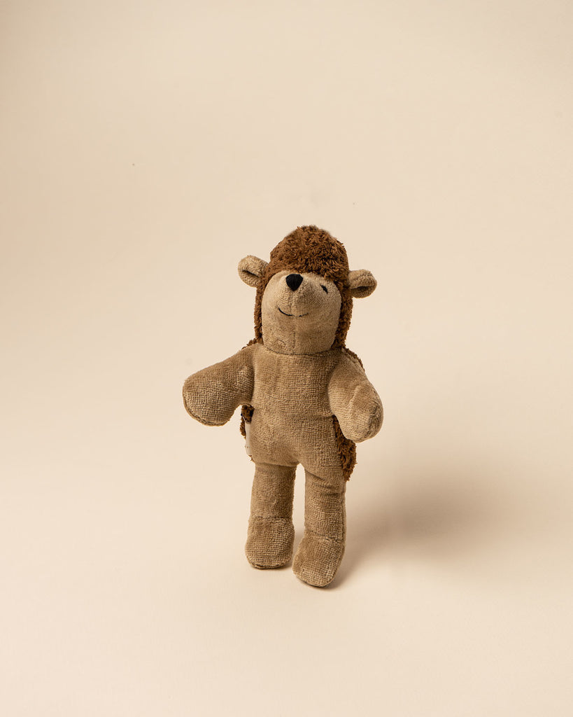 A well-loved brown and tan Senger Naturwelt stuffed hedgehog, handmade in Germany, stands against a plain, light beige background. The toy shows signs of wear, suggesting it is cherished.