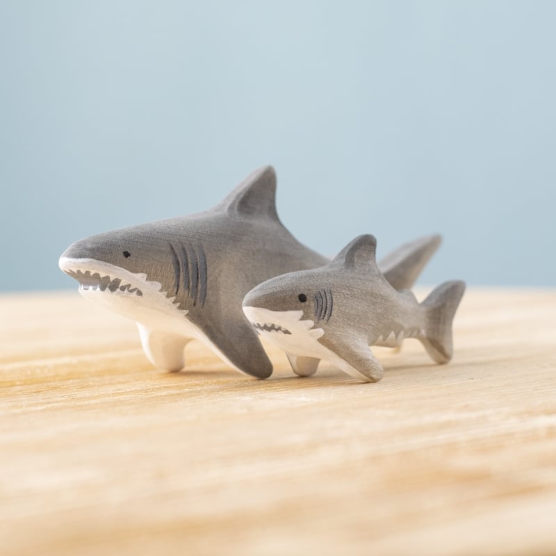 Two Bumbu Handmade Wooden Shark Family Set figurines, one large and one small, are displayed side by side on a light wooden surface against a plain blue background.