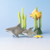 Two Bumbu Handmade Wooden Shark Family Sets, a yellow starfish, green seaweed, and an orange fish are arranged on a light blue background, creating an underwater Montessori toy scene.