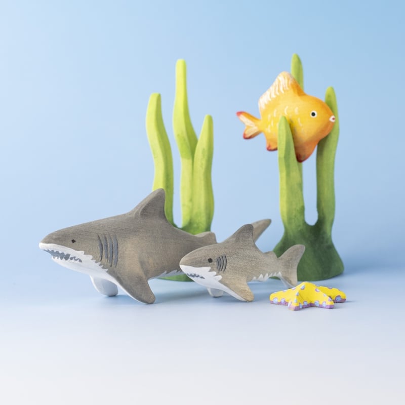 Two Bumbu Handmade Wooden Shark Family Sets, a yellow starfish, green seaweed, and an orange fish are arranged on a light blue background, creating an underwater Montessori toy scene.