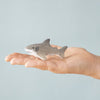 A person holds a small, gray and white Bumbu Handmade Wooden Shark Family Set toy in their palm against a light blue background—ideal for Montessori toy enthusiasts.