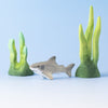 A shark toy from the Bumbu Handmade Wooden Shark Family Set sits between two green, coral-like plants on a light blue background, creating a playful, Montessori-inspired underwater scene.