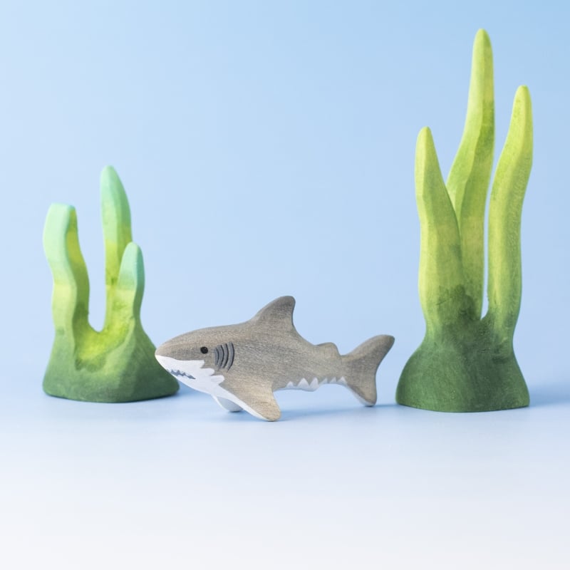 A shark toy from the Bumbu Handmade Wooden Shark Family Set sits between two green, coral-like plants on a light blue background, creating a playful, Montessori-inspired underwater scene.