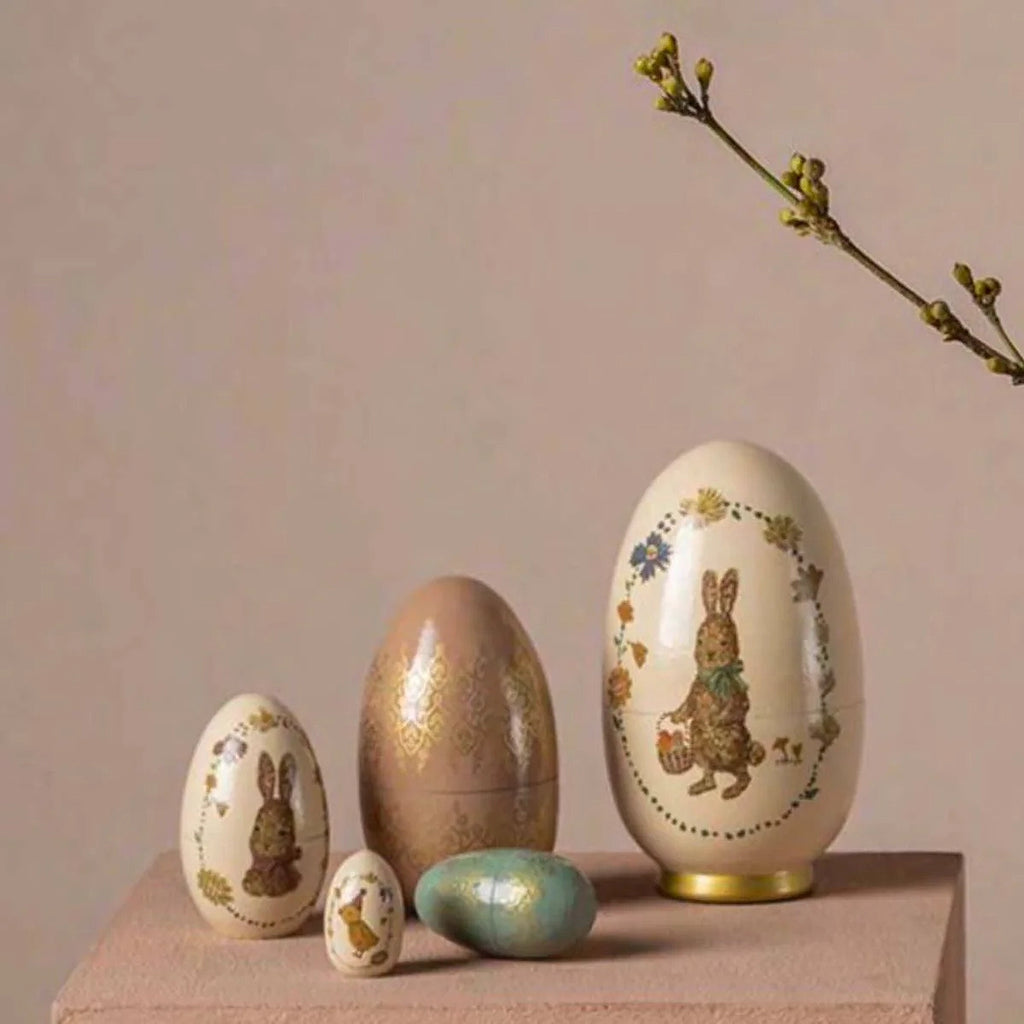 Four Maileg Easter Babushka Eggs, 5 pcs of varying sizes with intricate patterns and rabbit motifs, displayed elegantly against a neutral backdrop alongside a sprig of blooming flowers.