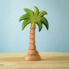 A Bumbu Wooden Small Palm Tree figurine, featuring a green leafy top and a brown trunk, is placed on a wooden surface. This charming toy, handcrafted from sustainable wood, sits against a light blue background, creating a simple and serene scene perfect for imaginative play.