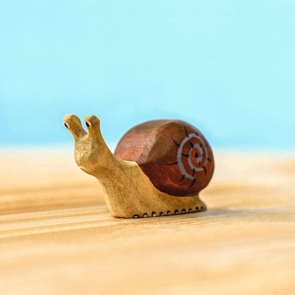 The Bumbu Handmade Wooden Snail, featuring a brown shell with a white spiral, is displayed on a light wooden surface with a blue background.