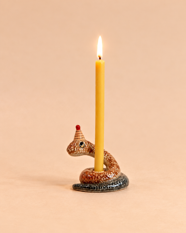 A yellow candle, lit and upright, is held by the coiled ceramic Snake Cake Topper—handcrafted with a small red party hat—set against a plain beige background.