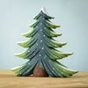 The Bumbu Handmade Wooden Snowy Fir Tree, Large, is an eco-friendly wooden toy painted green with white-tipped branches and small white dots to resemble snow, displayed on a light wooden surface against a pale blue background.
