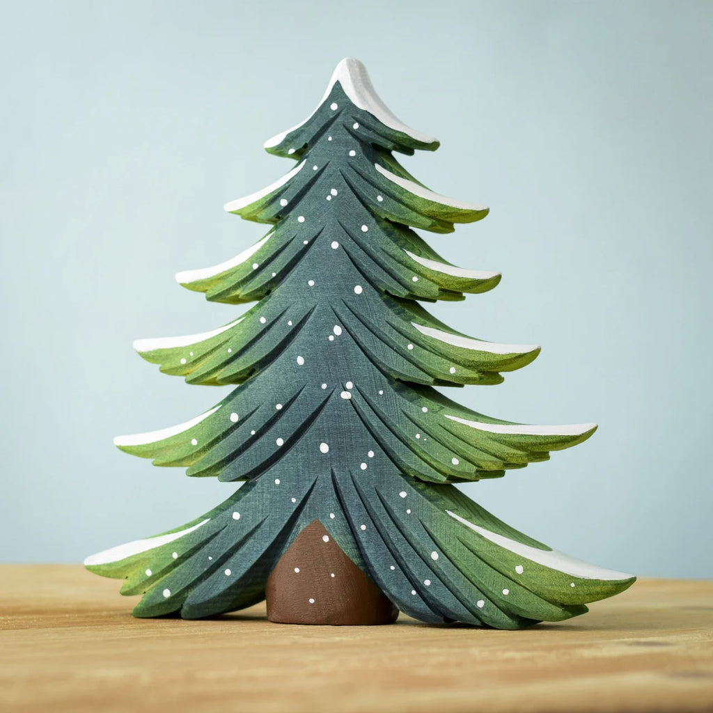 The Bumbu Handmade Wooden Snowy Fir Tree, Large, is an eco-friendly wooden toy painted green with white-tipped branches and small white dots to resemble snow, displayed on a light wooden surface against a pale blue background.