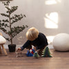 A young child plays on the floor with an eco-friendly wooden toy reindeer, a Santa figure, and the Bumbu Handmade Wooden Snowy Fir Tree (Large), near a small pine tree and a furry stool in a sunlit room.