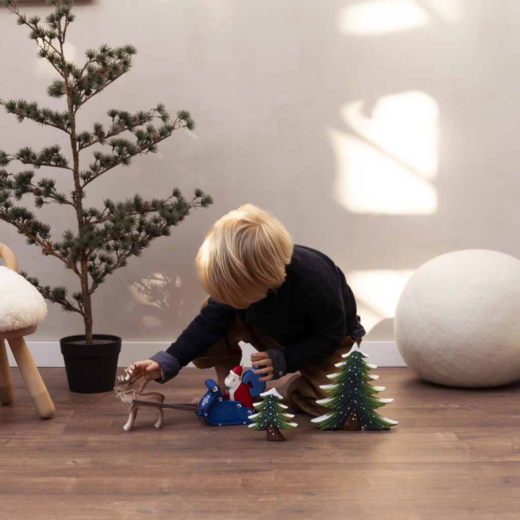 A young child plays on the floor with an eco-friendly wooden toy reindeer, a Santa figure, and the Bumbu Handmade Wooden Snowy Fir Tree (Large), near a small pine tree and a furry stool in a sunlit room.