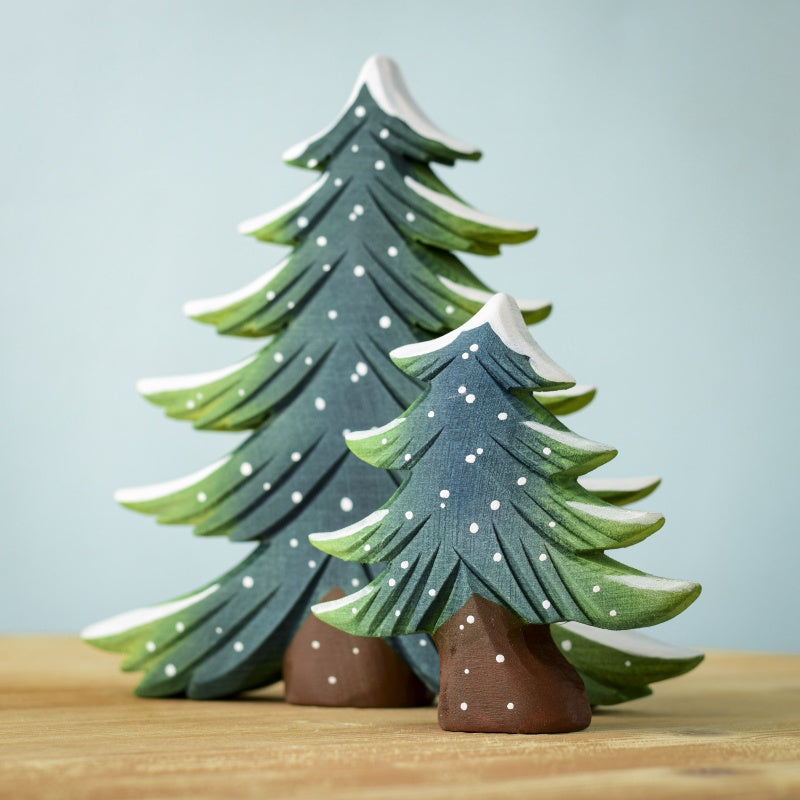 Two wooden figurines from the Snowy Fir Tree Set are elegantly painted in green with white snow accents, standing gracefully on a wooden surface. The larger tree stands behind the smaller one, creating a layered effect against the solid light blue background.