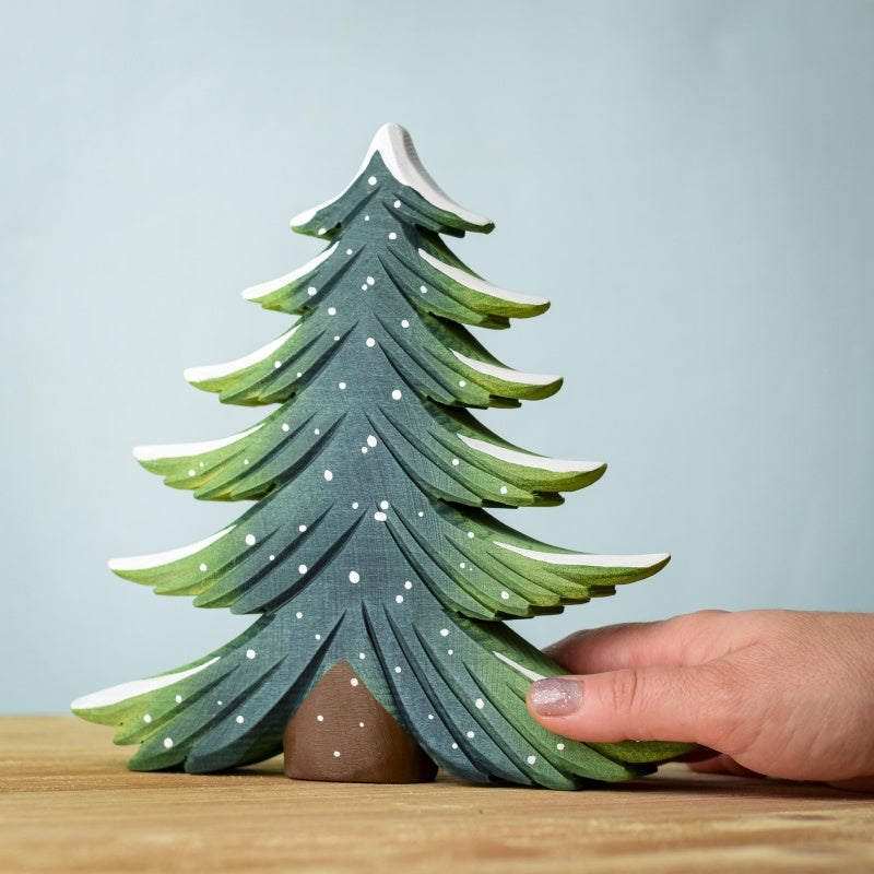 A hand gently holds a painted wooden pine tree from the Snowy Fir Tree Set, featuring snow accents. It's displayed on a wooden surface against a light blue backdrop, capturing the simplicity and natural beauty inspired by Waldorf designs.