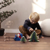 A young child with blonde hair is sitting on the floor, joyfully playing with the Snowy Fir Tree Set, which includes Christmas-themed decorations made from sustainably sourced wood. There are toy trees, a small Santa figure, and a reindeer next to him. A potted pine plant stands in the background.