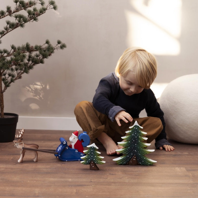A young child with blonde hair is sitting on the floor, joyfully playing with the Snowy Fir Tree Set, which includes Christmas-themed decorations made from sustainably sourced wood. There are toy trees, a small Santa figure, and a reindeer next to him. A potted pine plant stands in the background.
