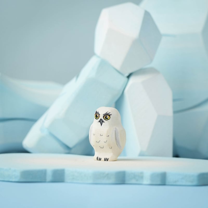 A small, white Bumbu Handmade Wooden Snowy Owl from the Wilderness Set with yellow eyes stands on a light blue icy surface, framed by geometric, ice-like blue shapes in the background.