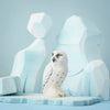 The Bumbu Handmade Wooden Snowy Owl from the Wilderness Set stands on a light blue surface, with geometric blue and white shapes in the background, evoking icy mountains or glaciers.
