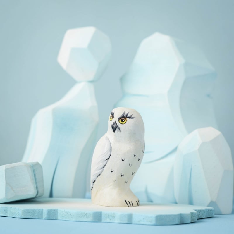 The Bumbu Handmade Wooden Snowy Owl from the Wilderness Set stands on a light blue surface, with geometric blue and white shapes in the background, evoking icy mountains or glaciers.