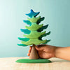 A hand holding an Extra Large Wooden Green Spruce Tree puzzle with layers in various shades of green and blue, painted with non-toxic paint, against a light blue background.