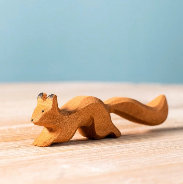The Bumbu Handmade Wooden Squirrel Running, a small handcrafted beechwood figurine, sits on a light wood surface with a soft blue background, ready to spark imaginative forest adventures.