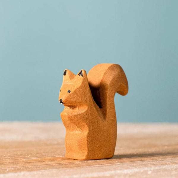 The Bumbu Handmade Wooden Squirrel Sitting, a small carved toy with a bushy tail, stands on a light wood surface against a plain blue background.
