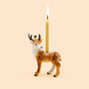 The Stag Cake Topper - Final Sale features a ceramic deer figurine adorned with white spots, delicately balancing a lit yellow candle on its back. Resembling a porcelain cake topper, the deer is crowned and set against a simple beige background.