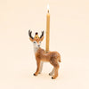 A handcrafted “Stag Cake Topper - Final Sale” ceramic deer figurine with a gold crown holds a lit candle on its back against a beige background.