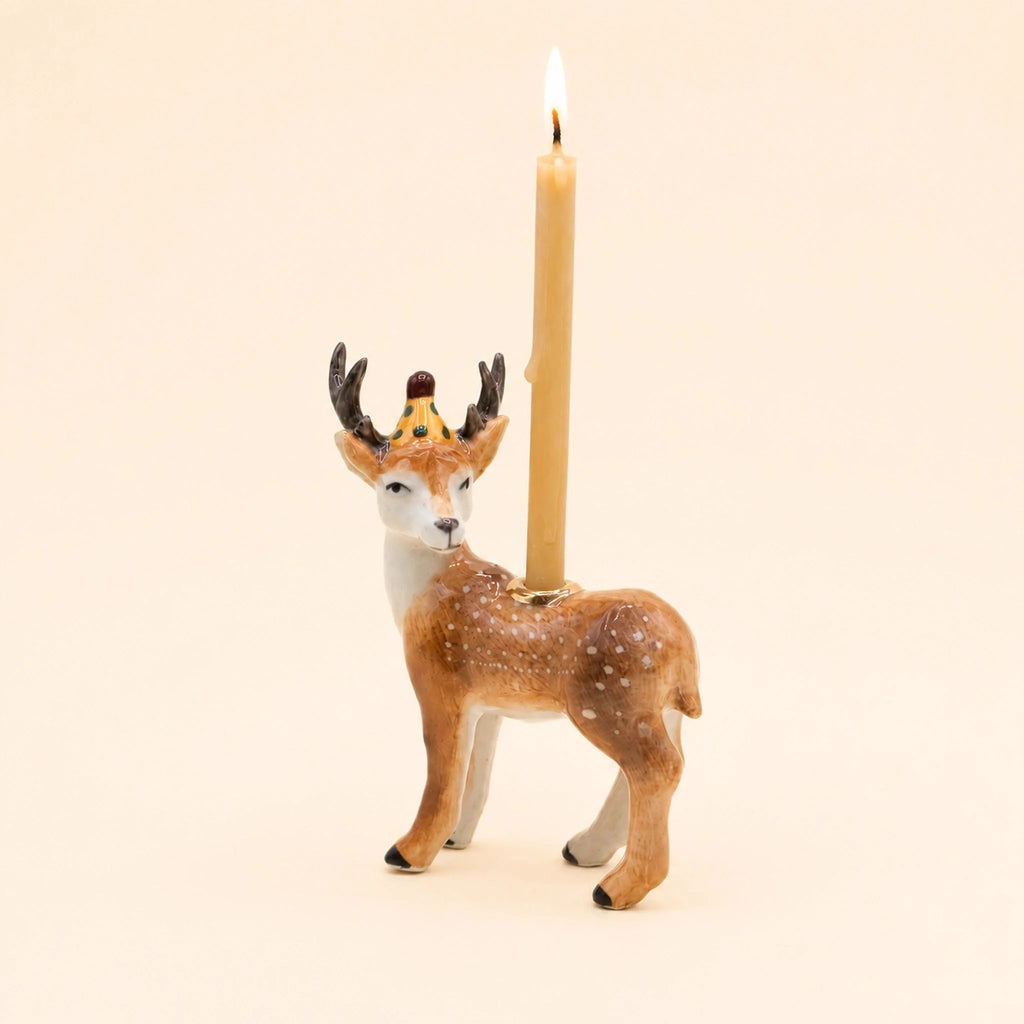 A handcrafted “Stag Cake Topper - Final Sale” ceramic deer figurine with a gold crown holds a lit candle on its back against a beige background.