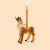 A handcrafted collectible, the Stag Cake Topper—a ceramic deer figurine adorned with a gold crown—holds a lit taper candle on its back, set against a plain beige background.