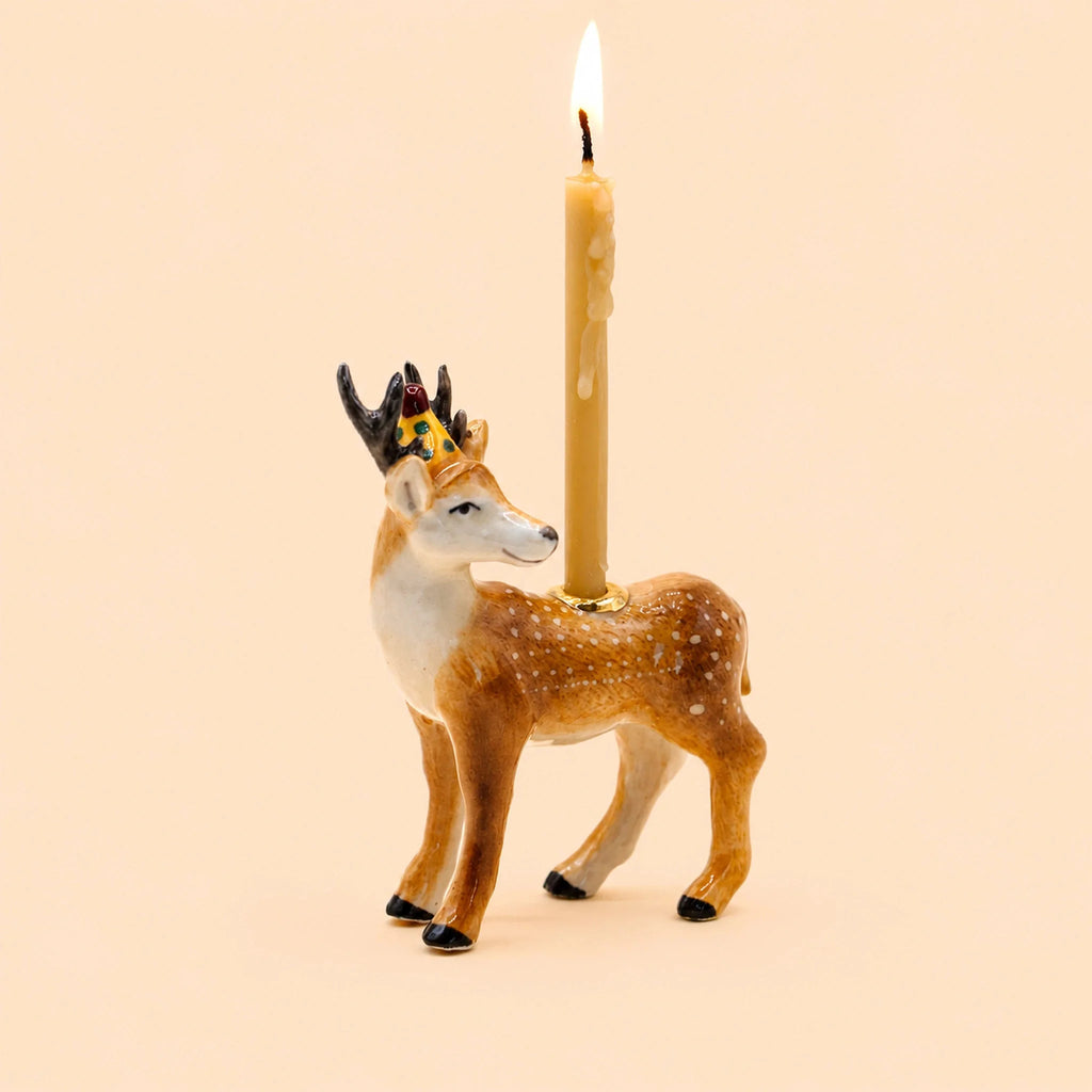 A handcrafted collectible, the Stag Cake Topper—a ceramic deer figurine adorned with a gold crown—holds a lit taper candle on its back, set against a plain beige background.