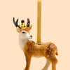 A handcrafted Stag Cake Topper, adorned with a yellow crown and colorful jewels, holds a slender yellow candle on its back against a plain beige background.
